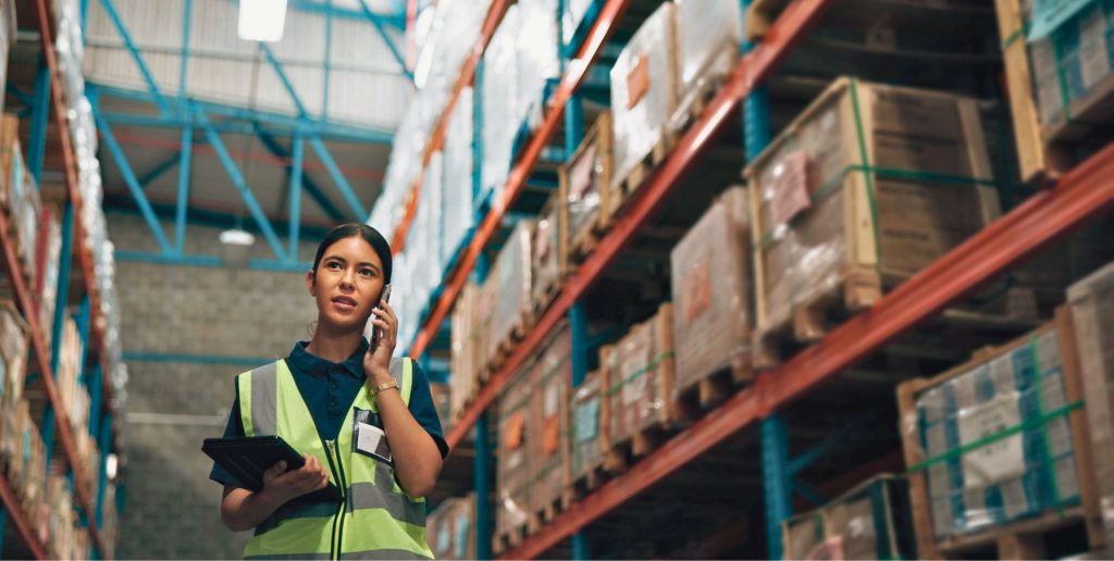 Woman in hi-vis vest oversees reliable transport in Melbourne warehouse, surrounded by industrial shelves and boxes.
