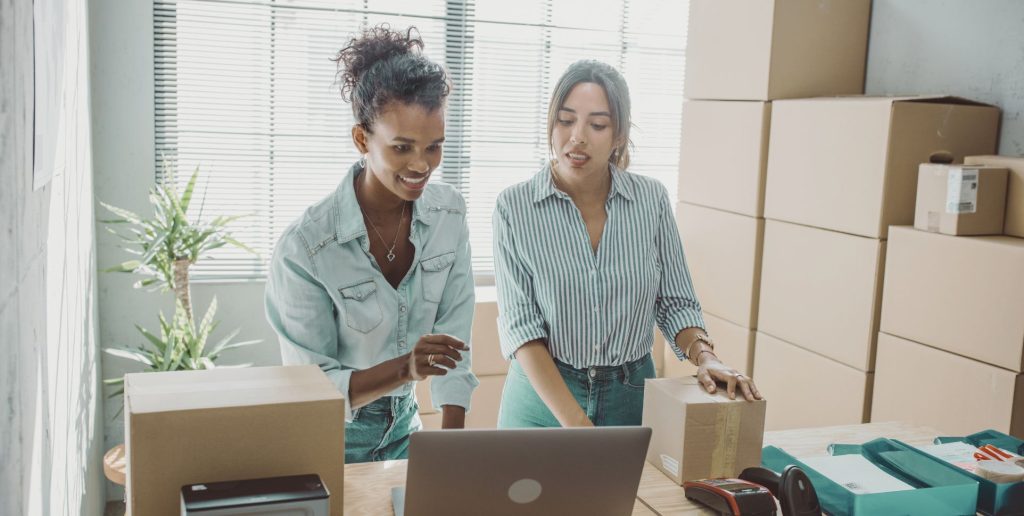 Two women prepare industrial consignments with cardboard boxes and laptop, showcasing Melbourne’s reliable transport services.